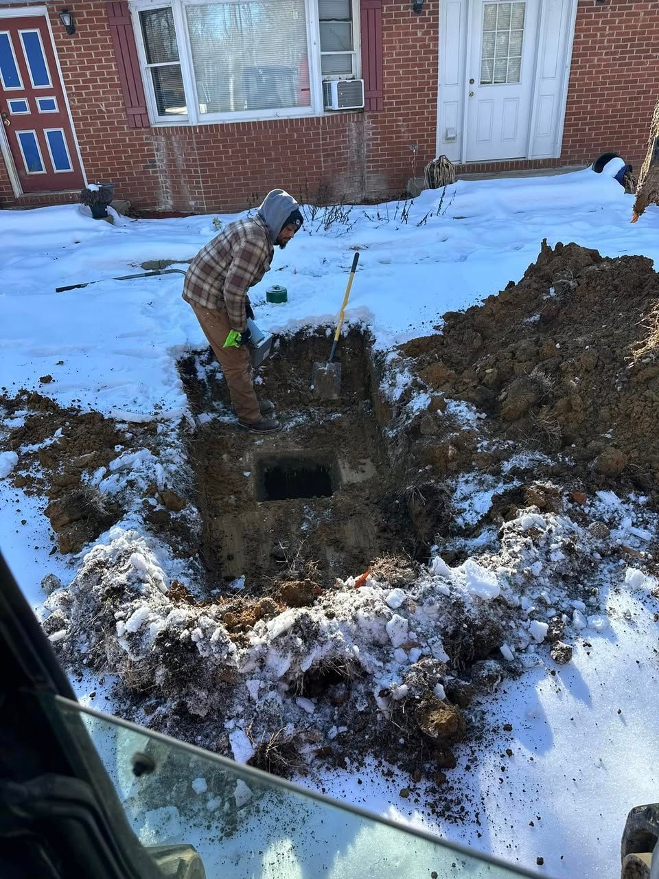 A man is digging a hole in the snow in front of a house for a septic system.