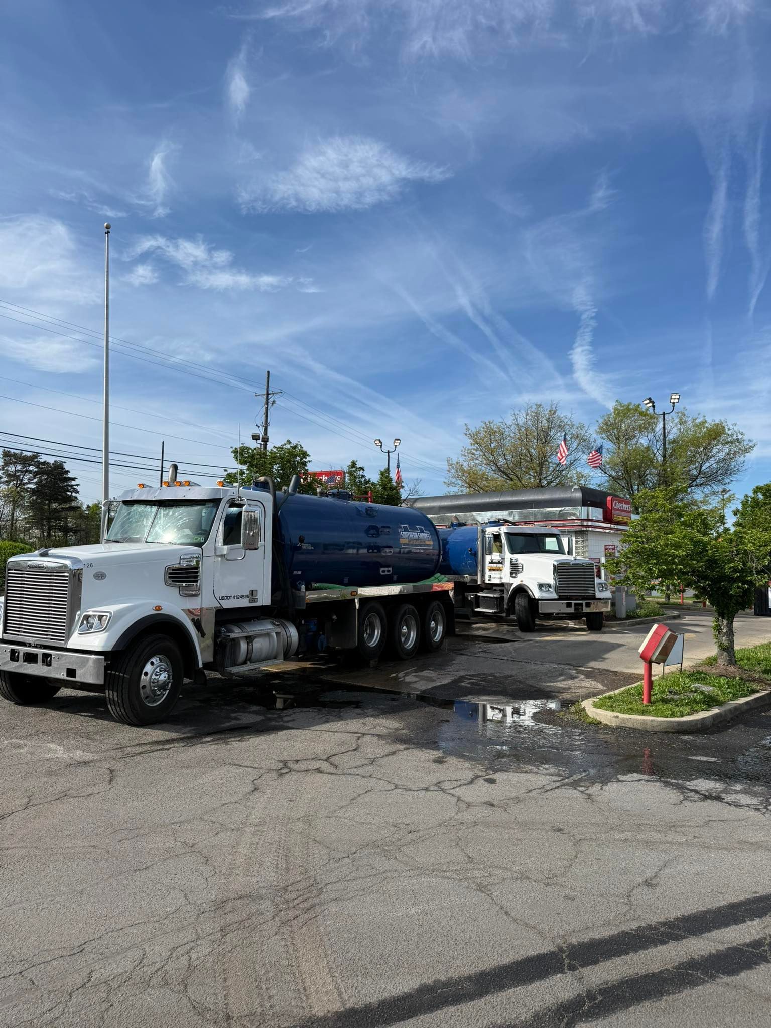 White tanker truck parked beside a curb under a bright blue sky with wispy clouds.