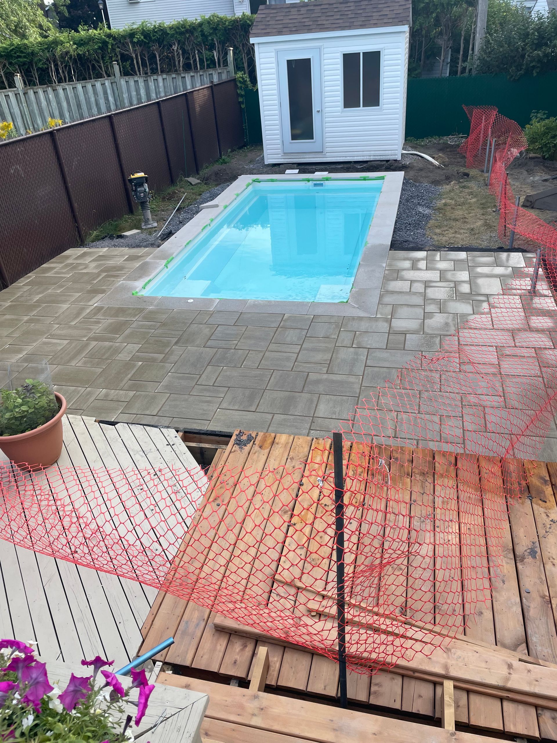 Cour arrière avec piscine, cabanon et terrasse en bois partiellement démontée; un filet de sécurité est présent.