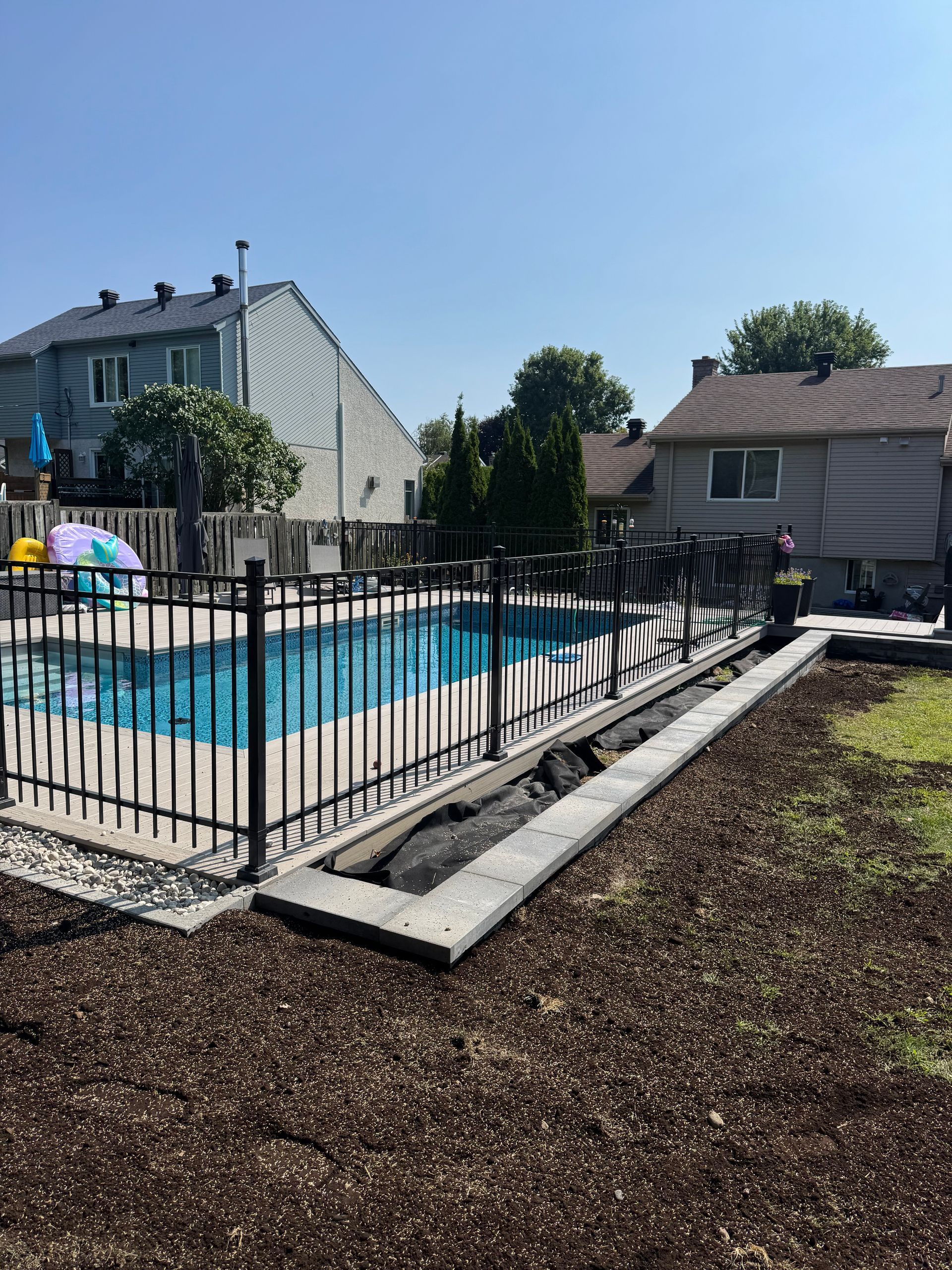 Piscine clôturée noire dans un jardin, entourée de paillis brun. Maisons en arrière-plan.
