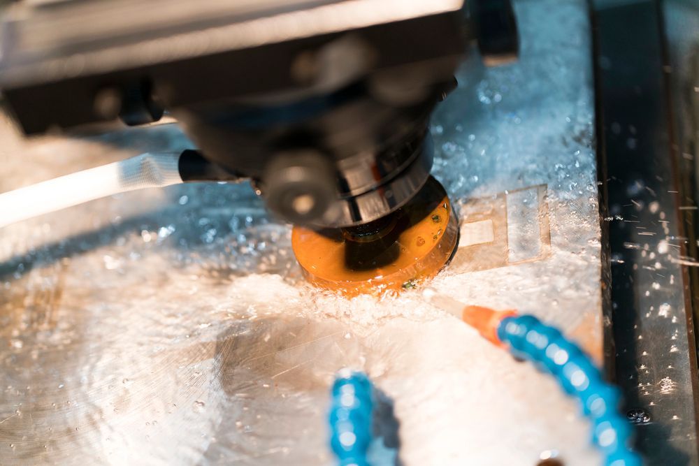 Fresadora CNC cortando metal, con refrigerante azul rociándose alrededor del área de la herramienta iluminada en naranja.