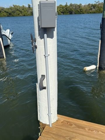 Gray electrical box and conduit on a dock post beside blue water and a wooden deck.