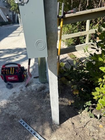 Electrical box on a post beside a sidewalk, with a tool bag and plants nearby.