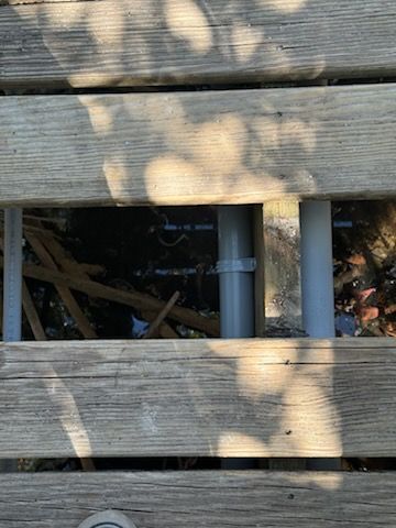 Wooden deck boards with a gray pipe visible underneath and dappled sunlight shadows