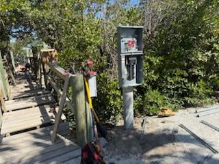 Electrical meter box beside a wooden walkway in a leafy outdoor area, with construction materials nearby