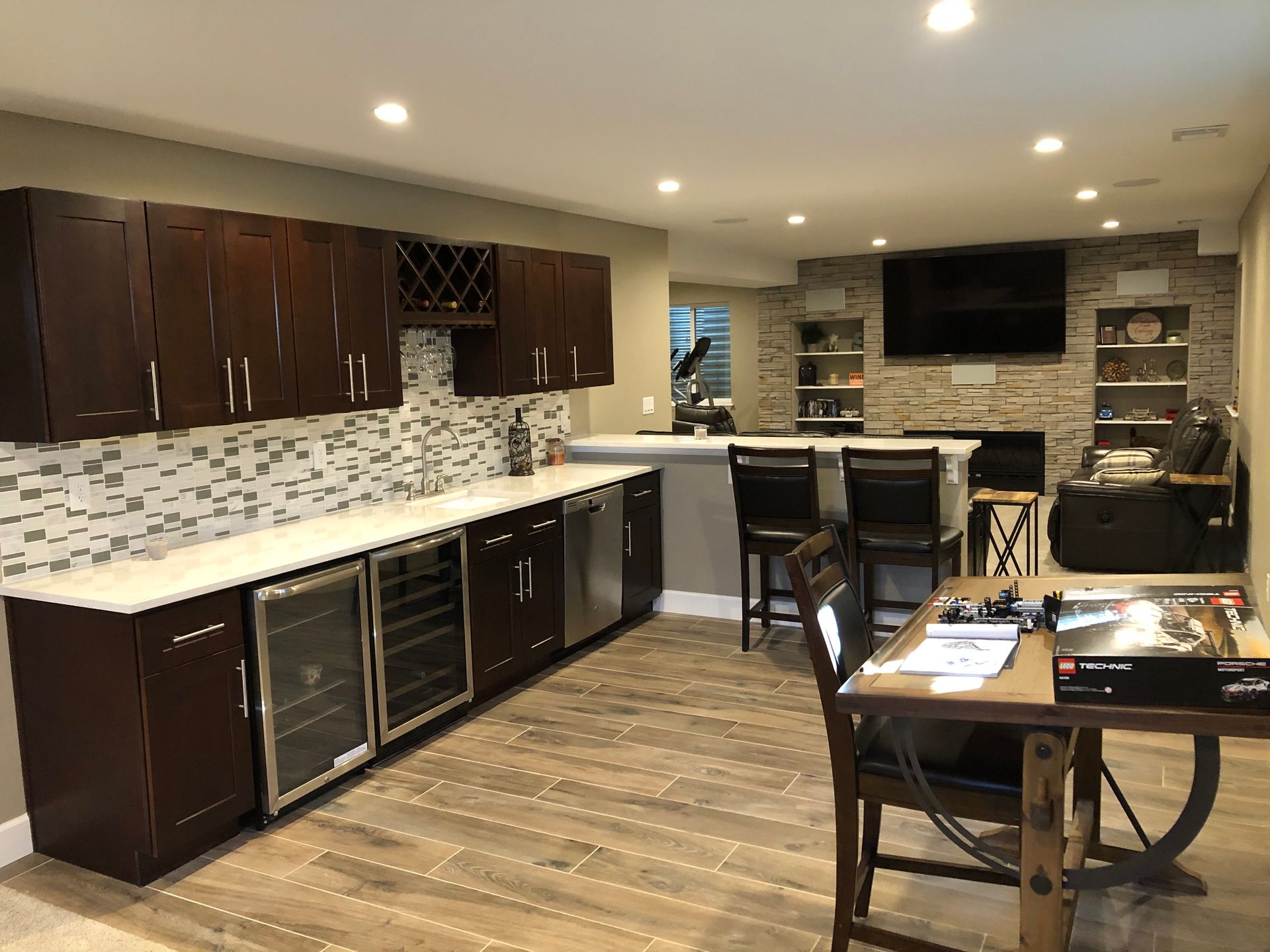 Basement bar area with dark wood cabinets, tile backsplash, and stone accent wall. Includes bar, wine fridge, seating, and entertainment system.