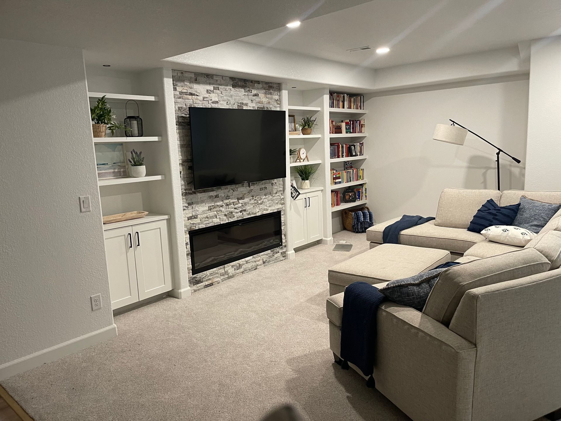 A cozy basement living room with a fireplace, TV, built-in shelves, and a sectional sofa on a light-colored carpet.