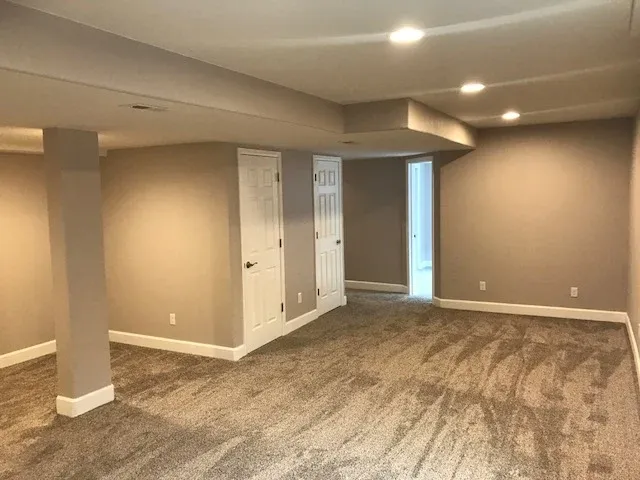 Finished basement with beige walls, tan carpet, white trim, and recessed lighting. Includes two support beams and two white doors.