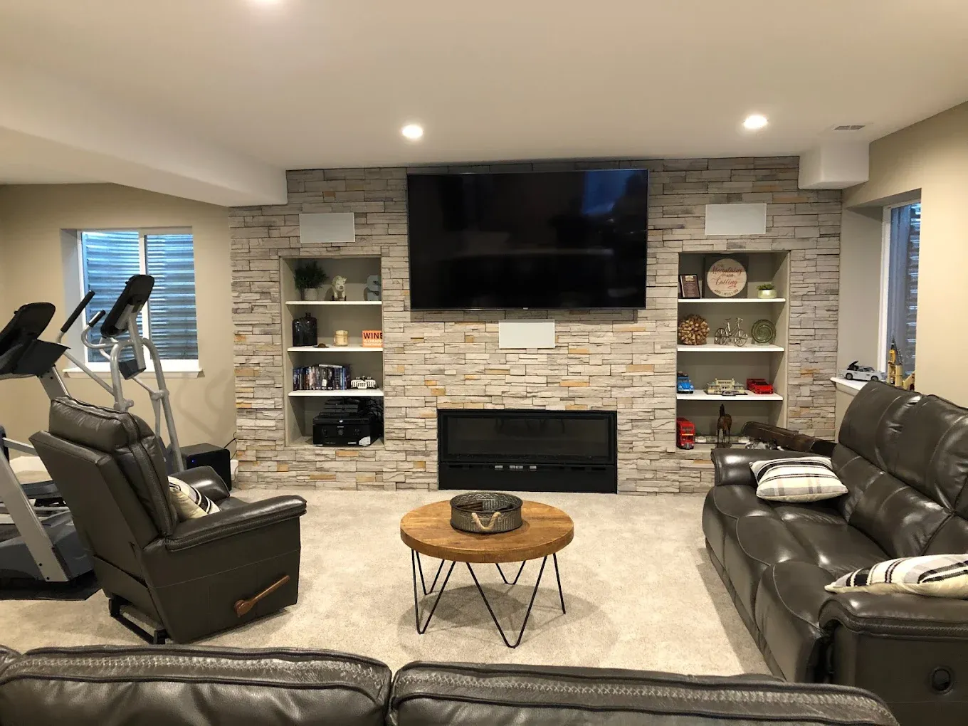 A finished basement with a stone accent wall housing a TV, fireplace, and built-in shelves, plus a treadmill and a leather couch.