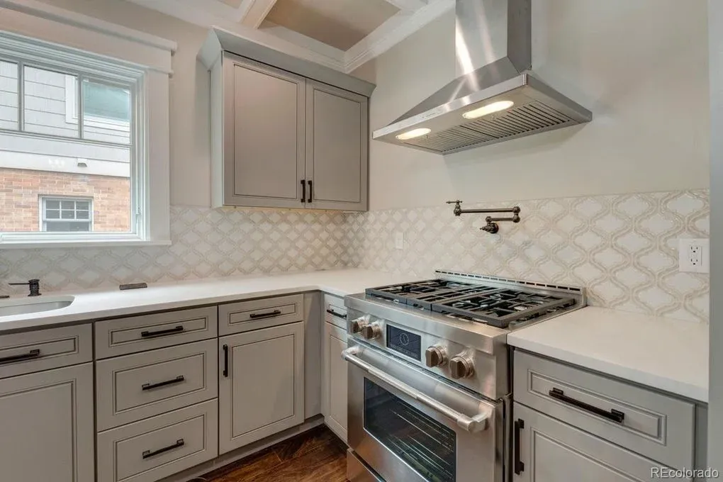 A modern kitchen with gray cabinets, white countertops, and a stainless steel range and hood. A white tile backsplash and a window are also present.