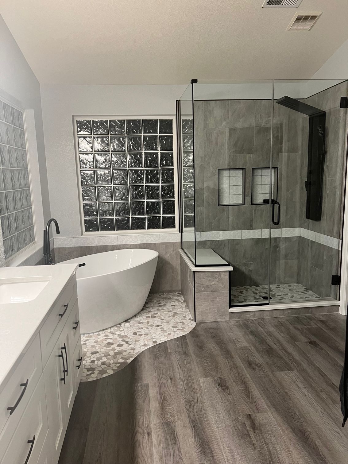 Modern bathroom with white tub, glass shower, grey tile, and light wood flooring.