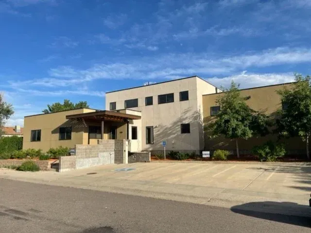 Two-story tan and white building with a covered entrance and parking area, under a blue sky.