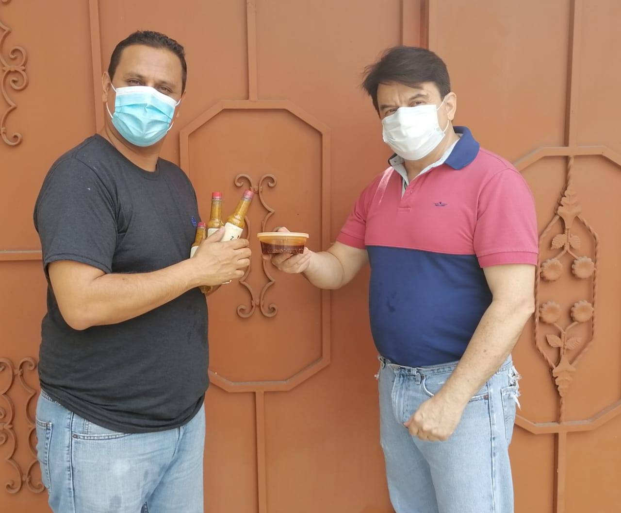 Two masked men clink drinks in front of a brown door and ornate gate.