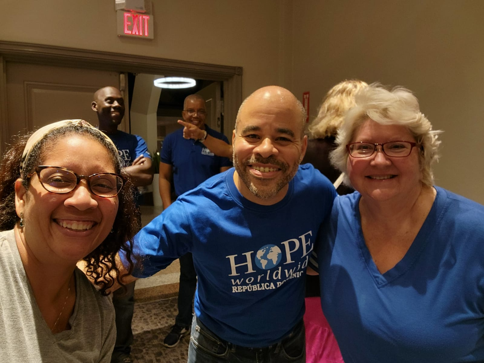 Three smiling people posing indoors at a gathering, wearing blue shirts; others stand in the background.