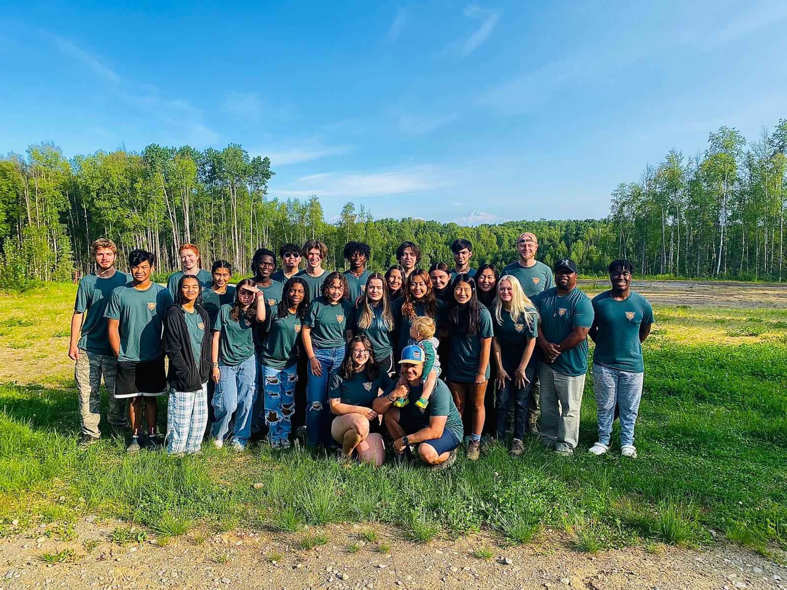Group of people in teal shirts posing outdoors on a sunny grassy field with trees behind them