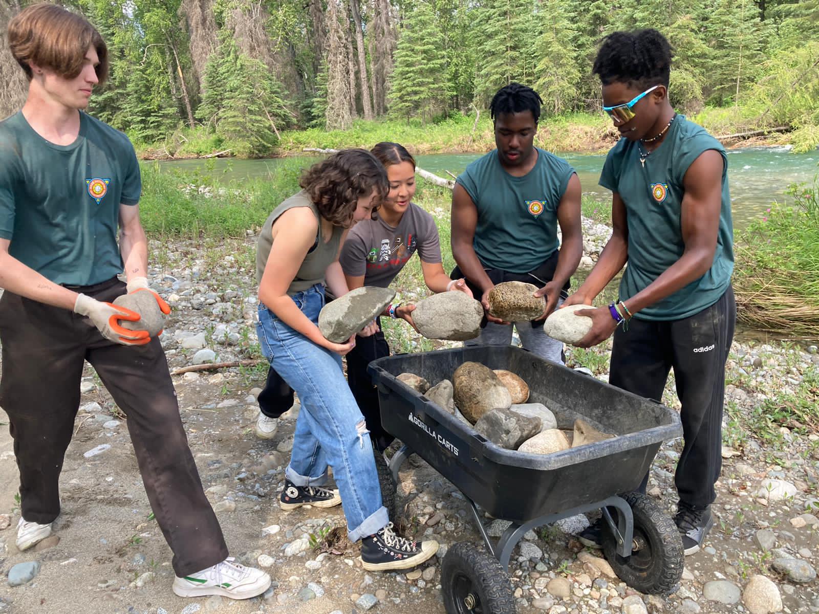 Four people outdoors load rocks into a black wheelbarrow beside a stream and trees.