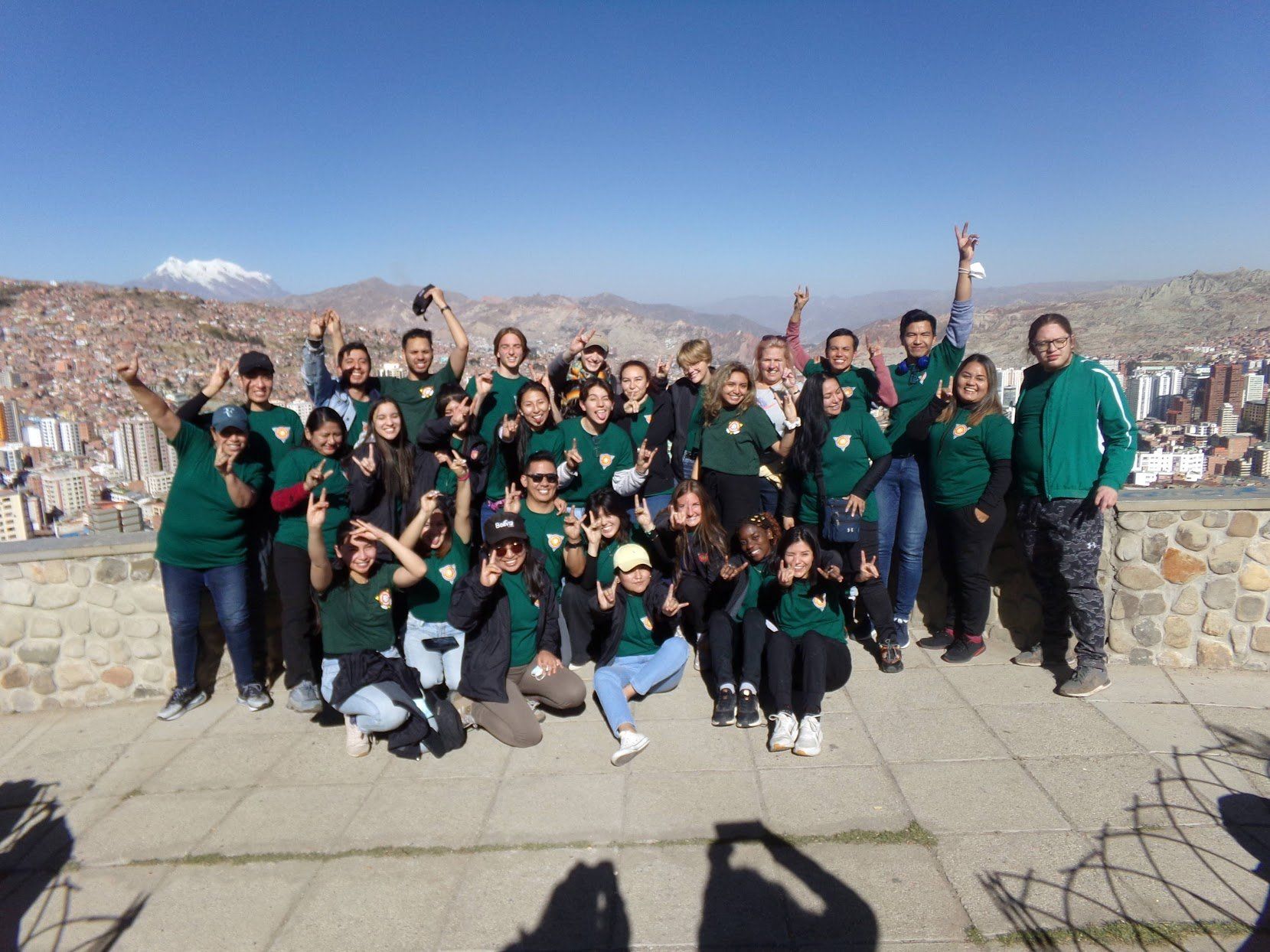Group of people in green shirts posing on a stone overlook with city and mountains behind them.