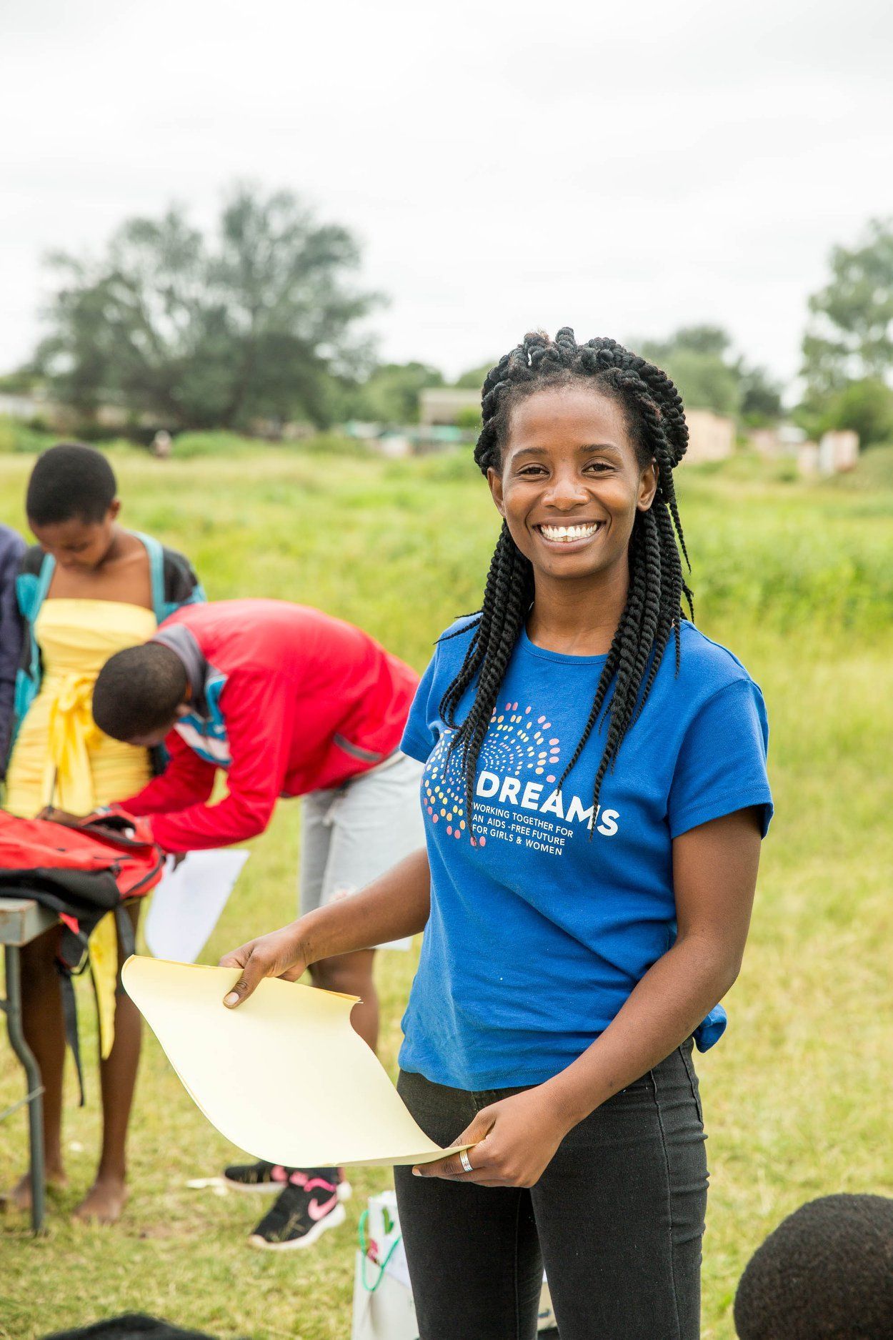 Smiling volunteer in blue shirt holding a yellow sheet outdoors while others work nearby