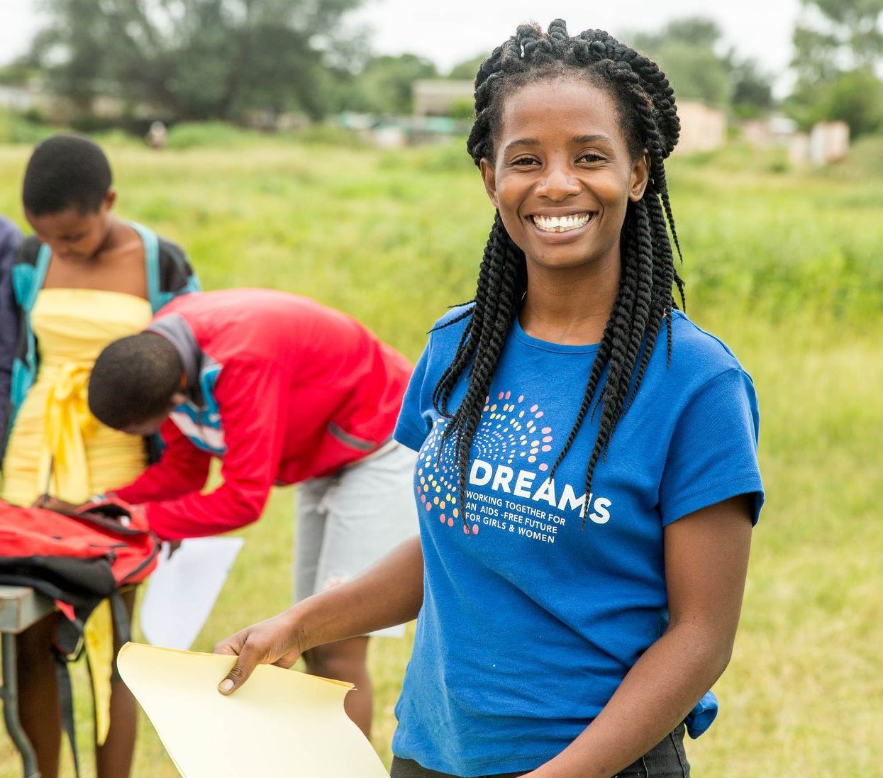 Smiling volunteer in blue shirt holding papers, with others packing supplies outdoors on a grassy field