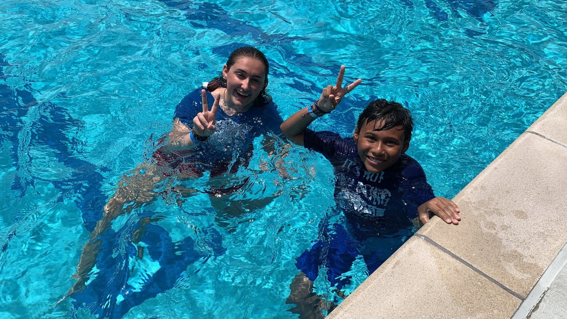Two people in dark shirts smiling in a bright blue swimming pool, one by the pool edge