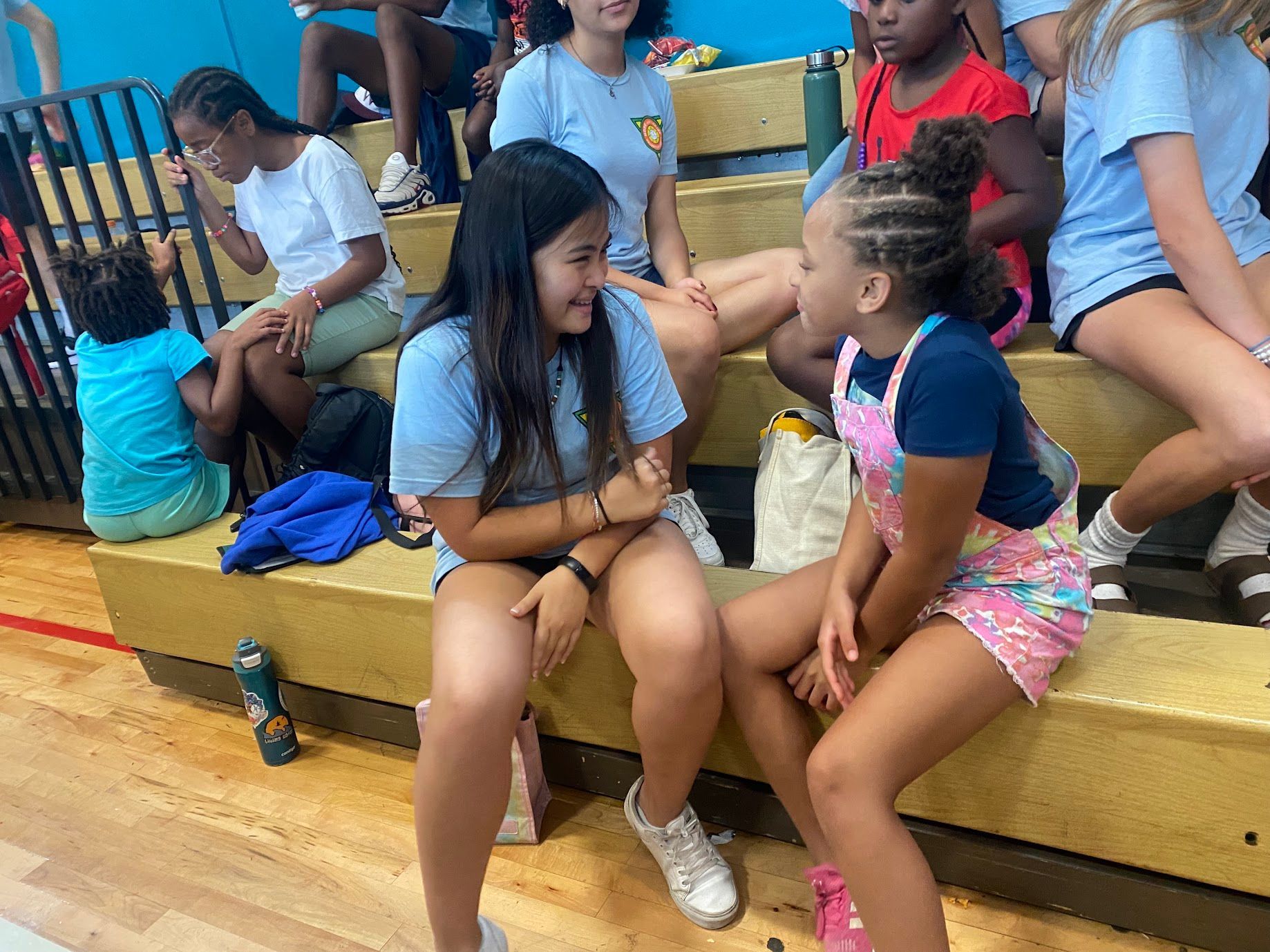 Two girls chatting on wooden bleachers at an outdoor sports event, with bags and spectators nearby
