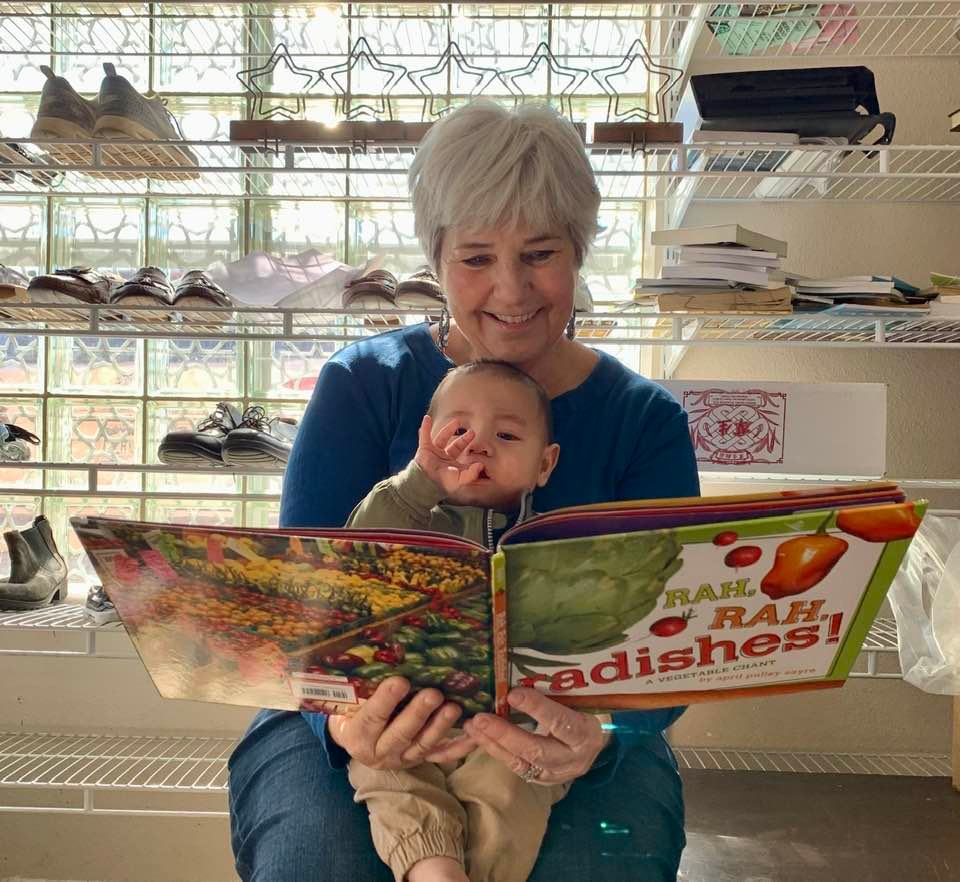 Person holding a baby while reading a colorful book in a bright room with shelves behind them