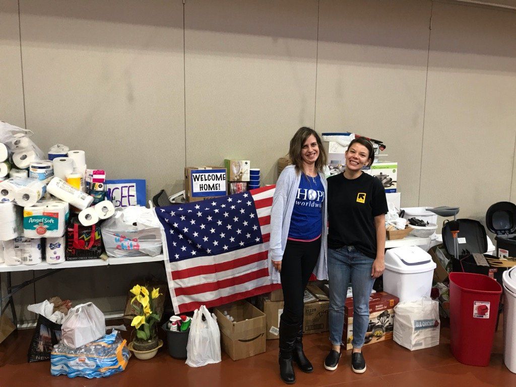 Two people standing in a cluttered room with an American flag and boxes behind them