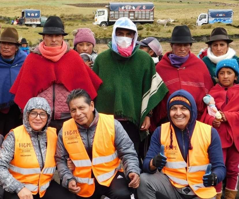 Group of people in colorful jackets and hats posing outdoors on a grassy field, with trucks in the background