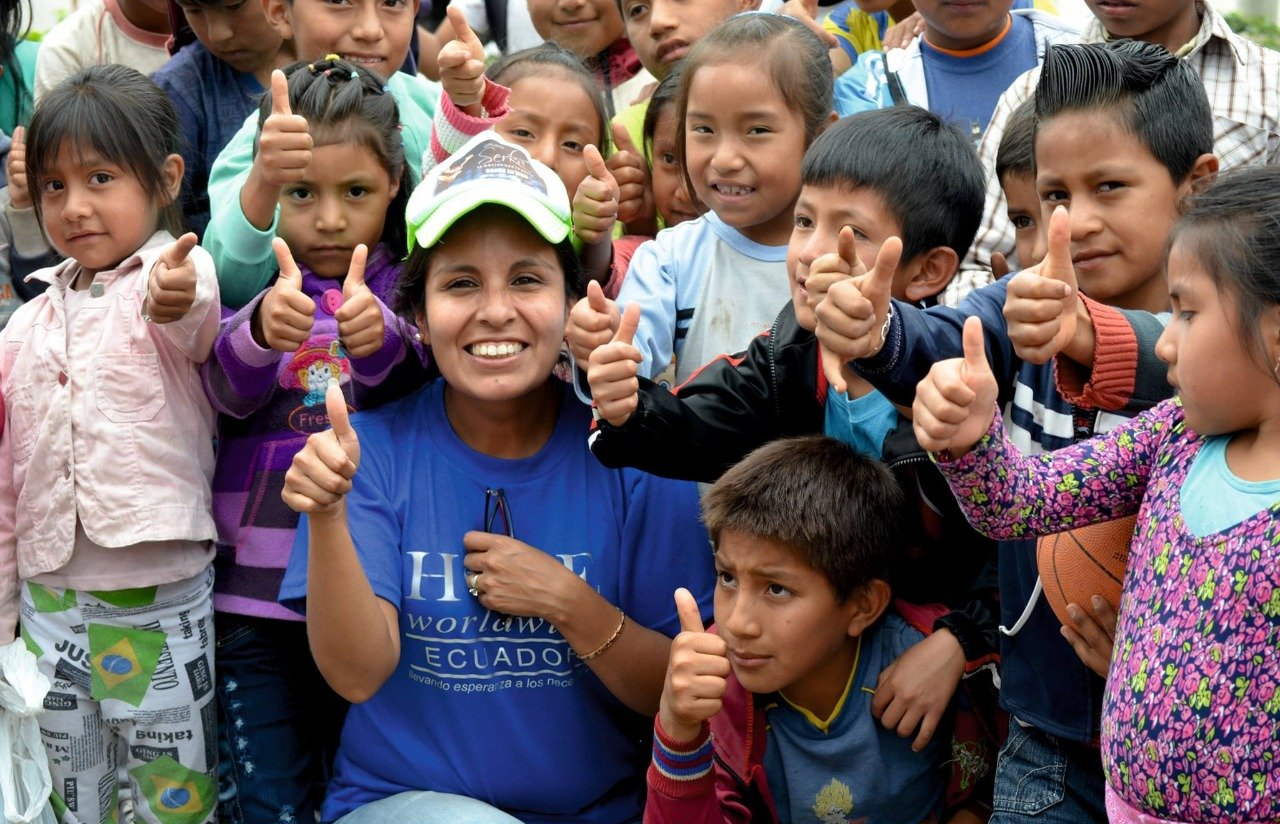 Group of smiling children outdoors, posing with thumbs up and peace signs.