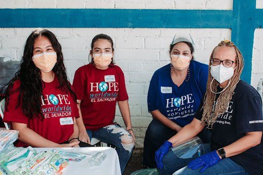 Four masked volunteers sit indoors beside a table, wearing matching red and blue shirts.