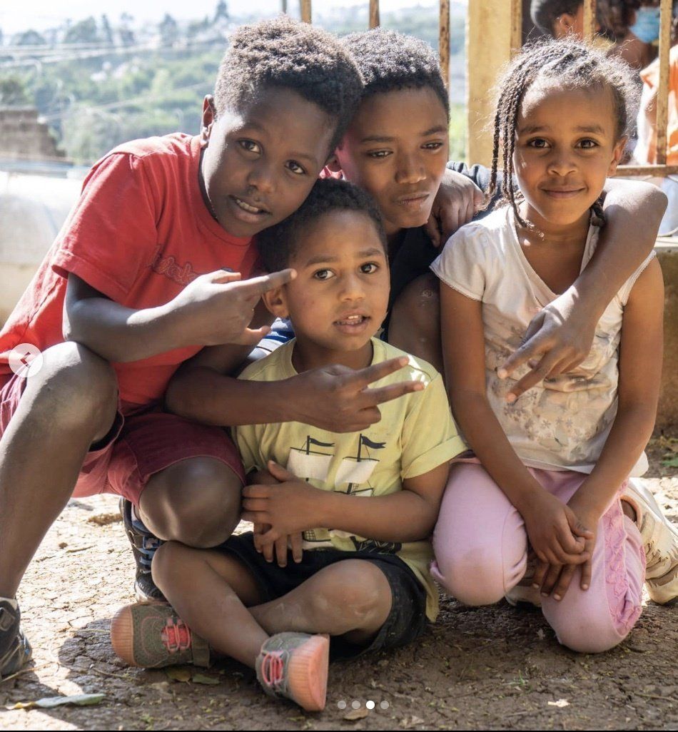 Four children crouch together outdoors, smiling at the camera in bright sunlight.