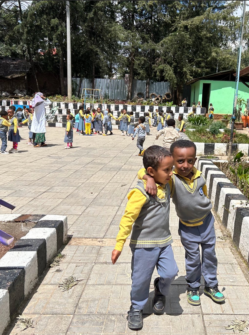 Two children in gray and yellow uniforms stand arm-in-arm in a school courtyard with other children behind them.