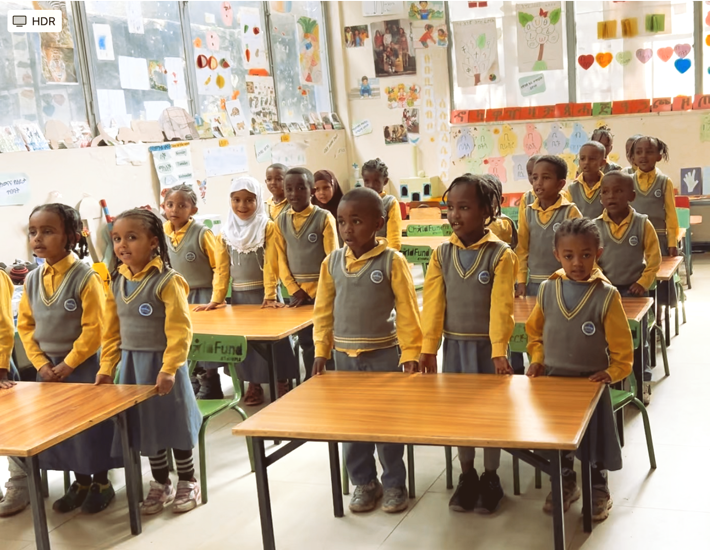 Children in matching yellow uniforms stand beside desks in a classroom.
