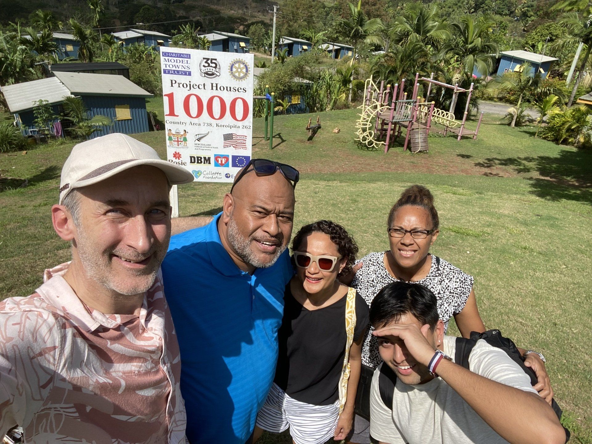 Five smiling people posing outdoors on a sunny lawn with a sign behind them.