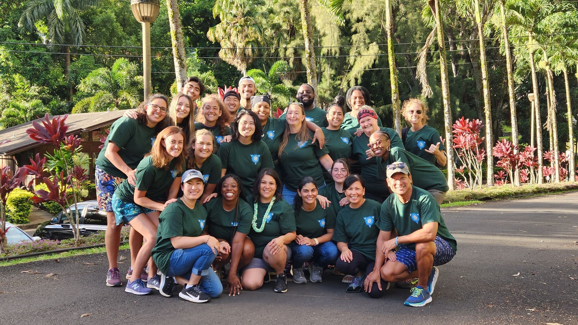 Group of people posing together outdoors in green shirts with tropical trees and flowers behind them