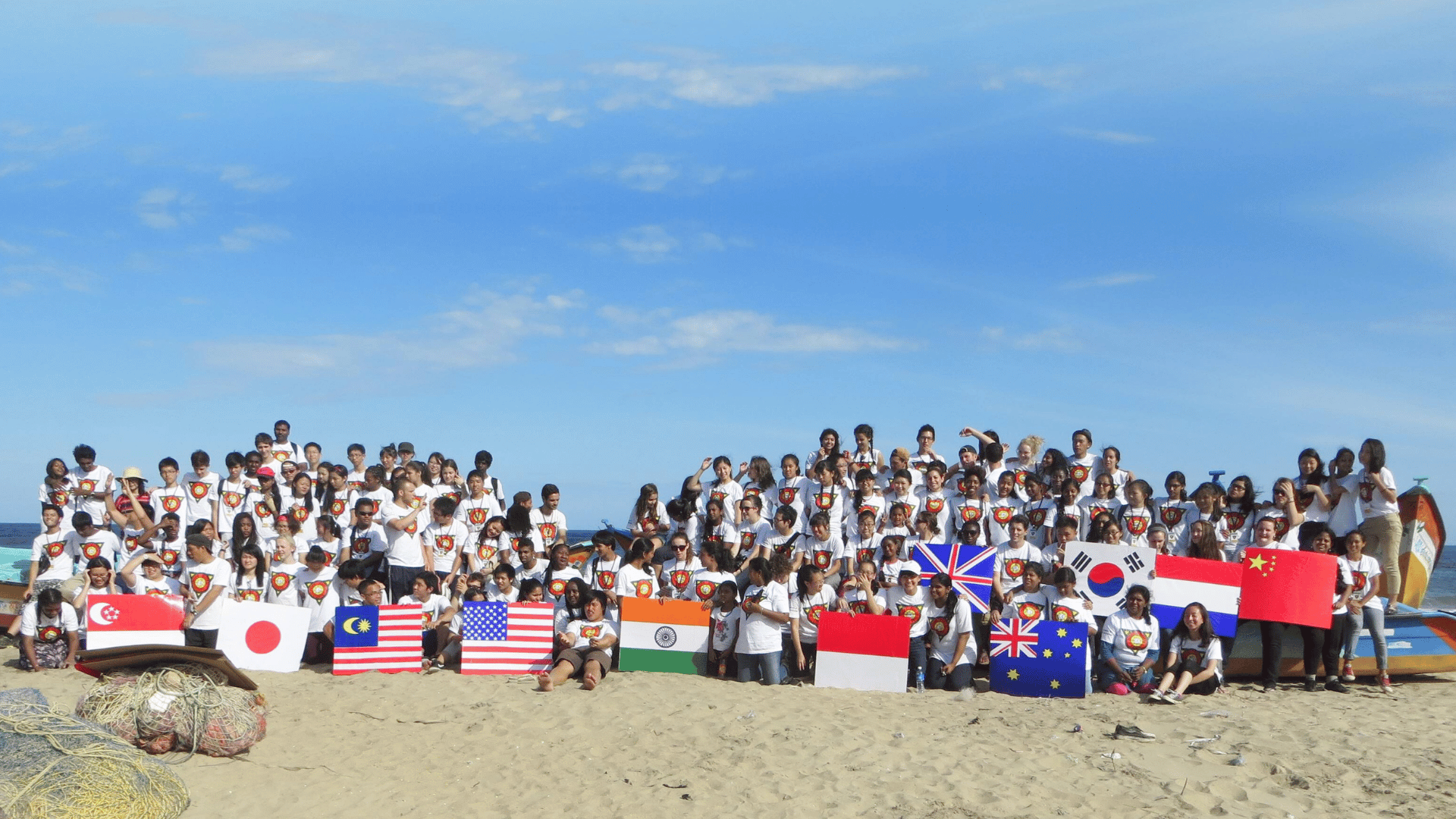 Large group posing on a beach behind a row of national flags under a blue sky