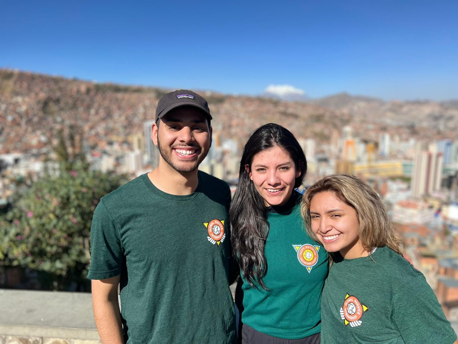 Three people in green shirts smiling outdoors with a city and mountains behind them