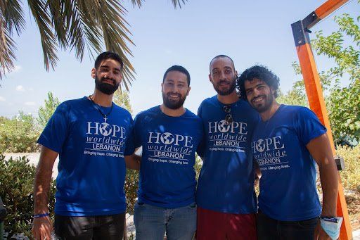 Four people in blue T-shirts stand together outdoors, smiling under palm trees.