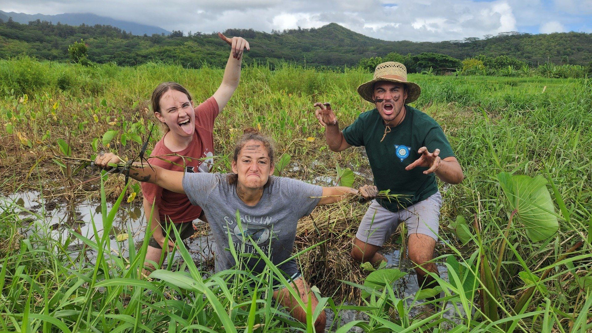 Three people posing in a muddy green field, smiling and raising their arms, with hills in the background.