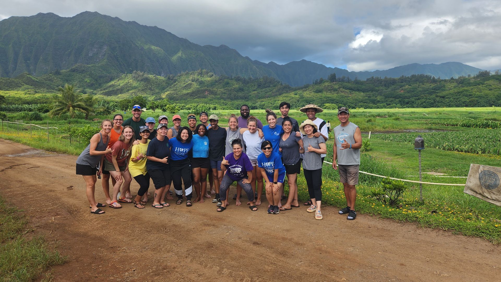 Group of people posing on a dirt road beside green fields with mountains and cloudy sky in the background
