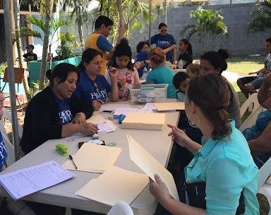 People gathered around tables outdoors, writing and discussing papers in a shaded patio area.