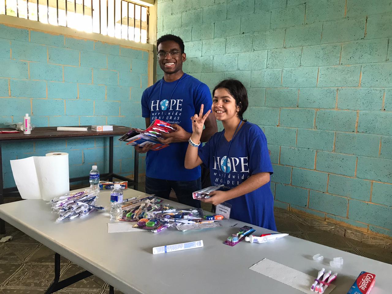 Two volunteers in blue shirts stand behind a table with wrapped items in a room with teal walls.