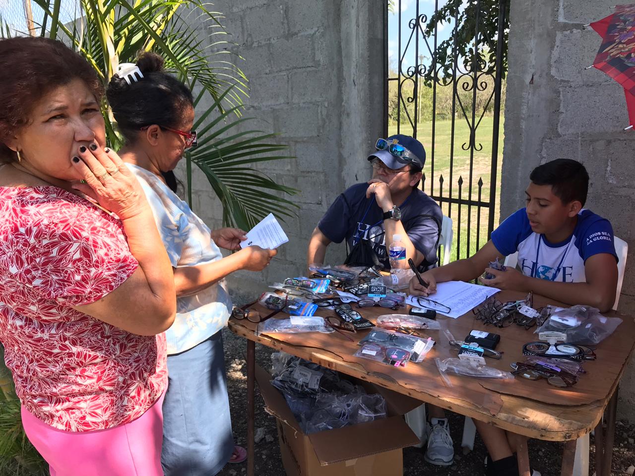 People at an outdoor table examining and sorting small items near a gate and concrete wall.