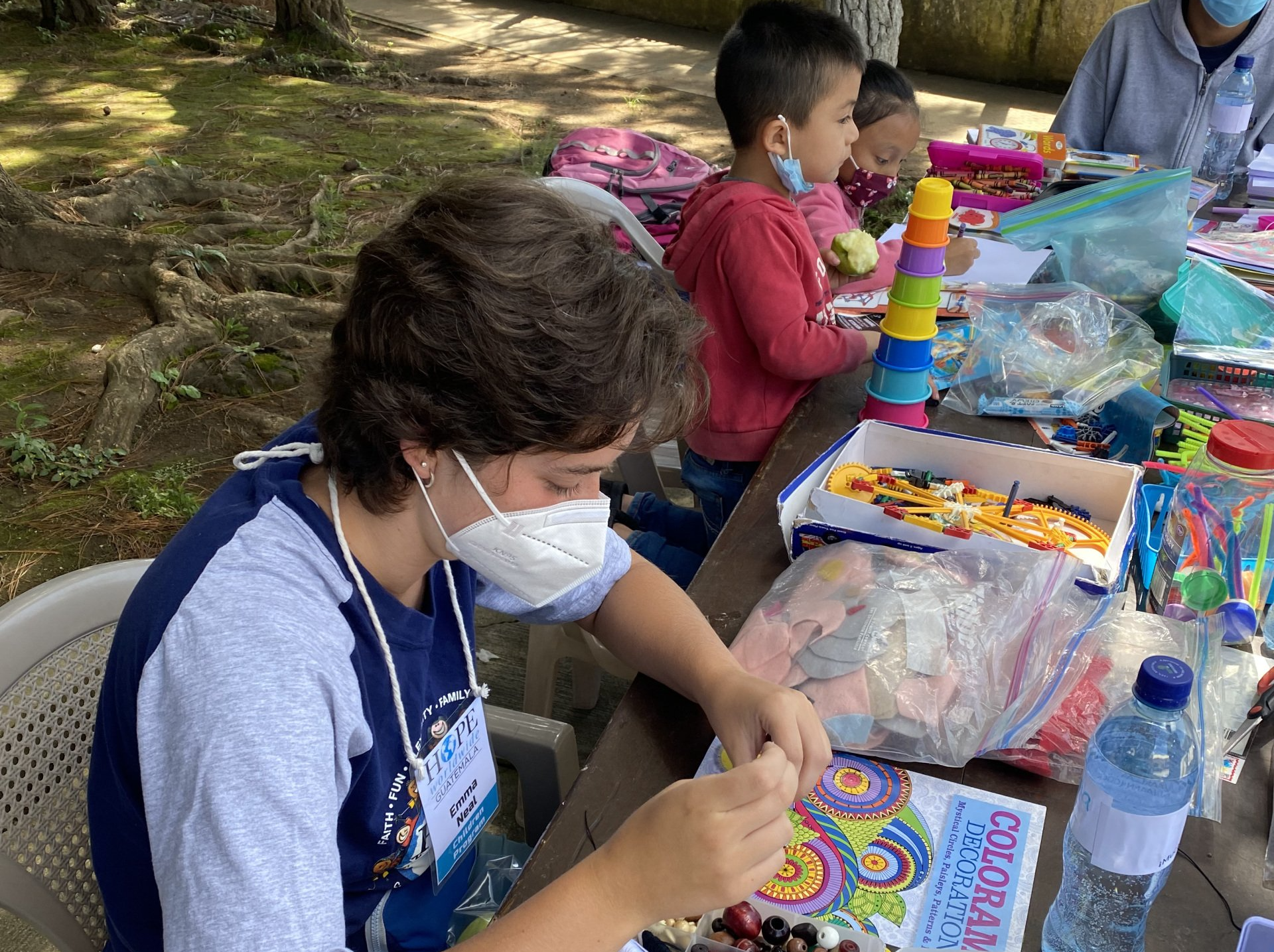 Masked child painting at an outdoor table with craft supplies and colorful toys