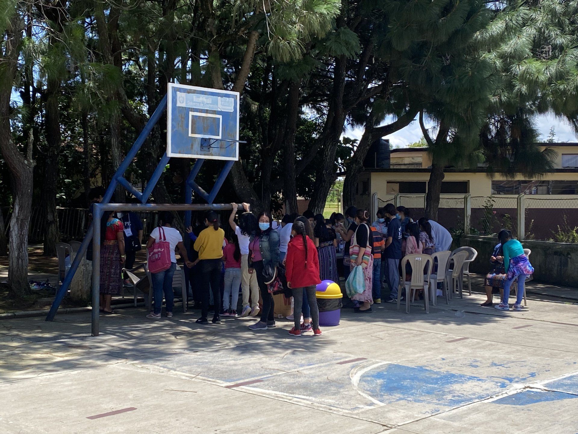 People gathered under a basketball hoop in a shaded outdoor court.