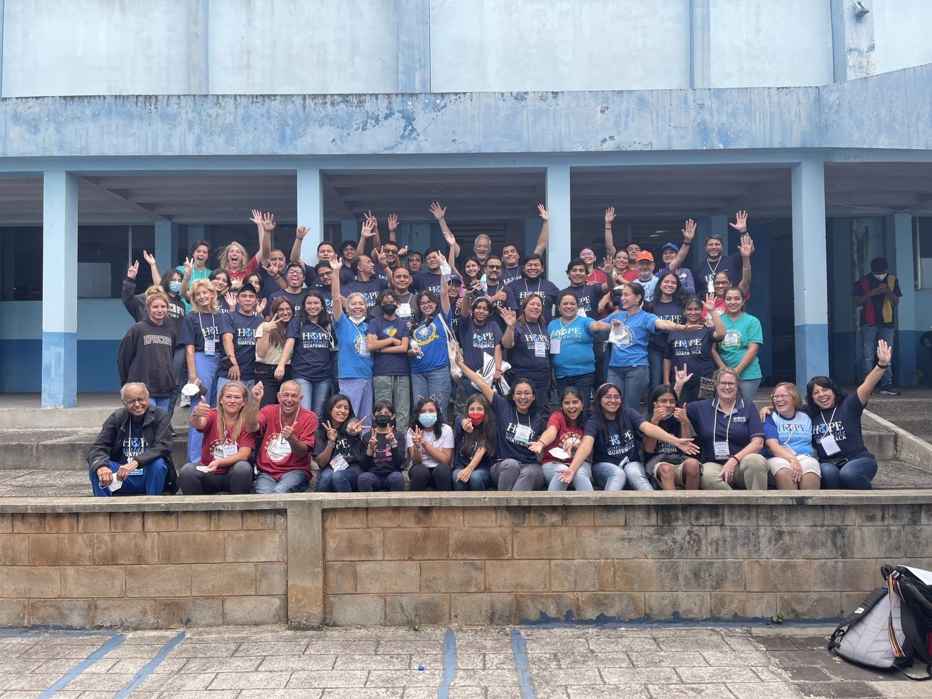 Large group posing on outdoor steps in front of a concrete building, many smiling and raising their hands