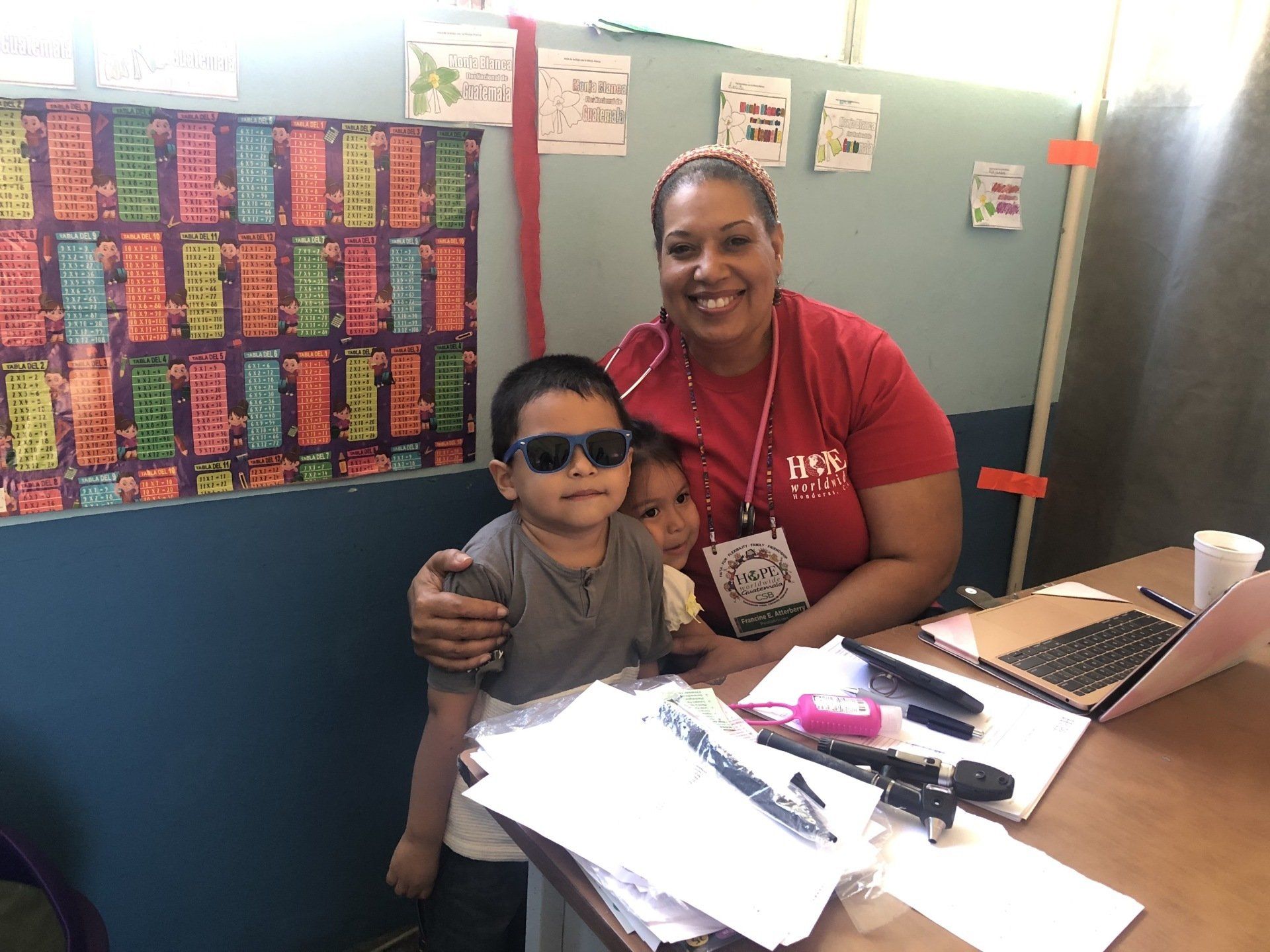 Woman with two children at a desk in a classroom, smiling beside a laptop and colorful wall display