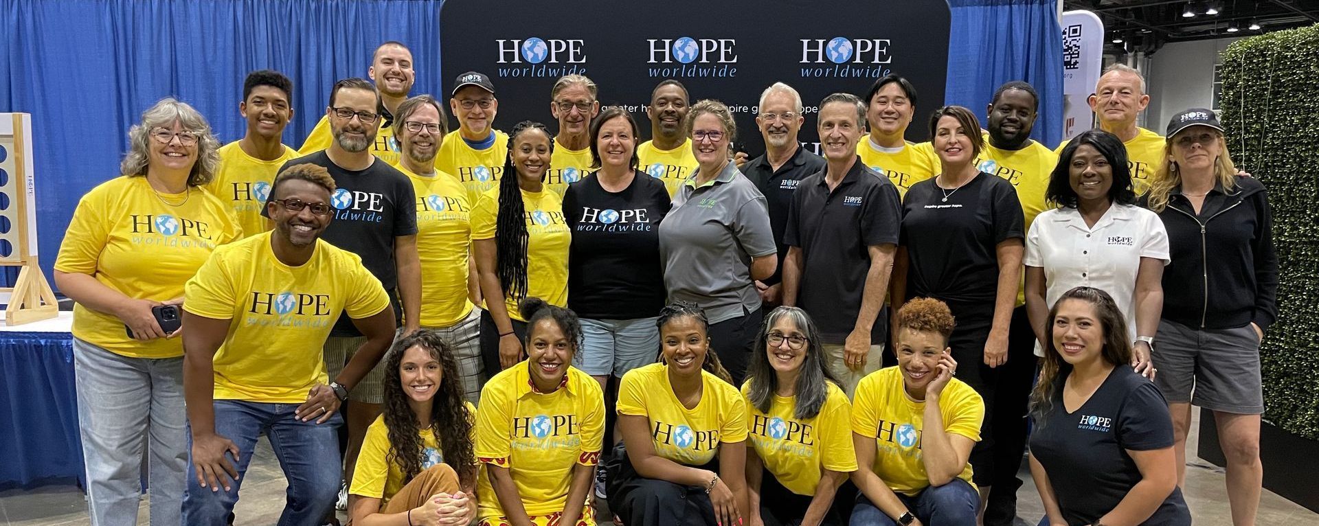 Group of volunteers posing indoors in front of a blue backdrop, many wearing yellow shirts and smiling.