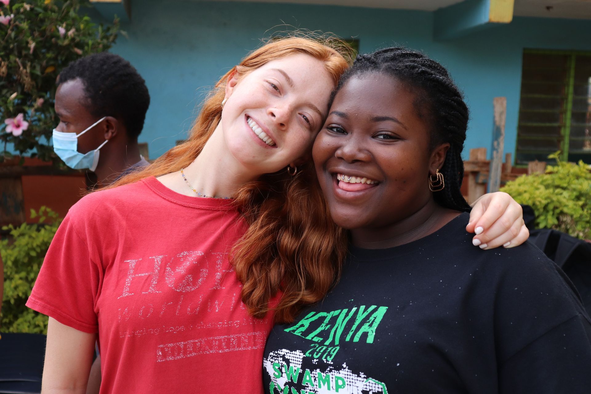 Two smiling people posing arm-in-arm outdoors, one in a red shirt and one in a black shirt.