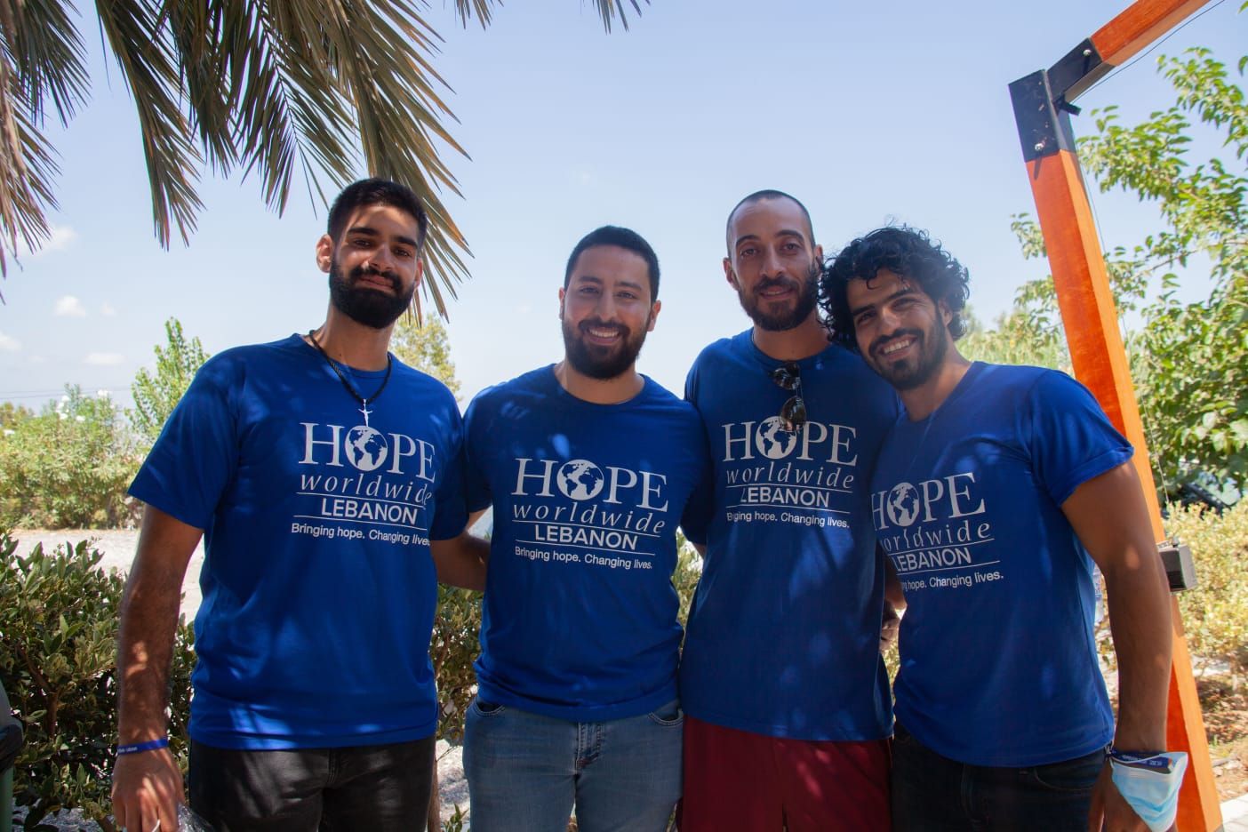 Four smiling people in blue “HOPE” T-shirts standing outdoors under palm trees.
